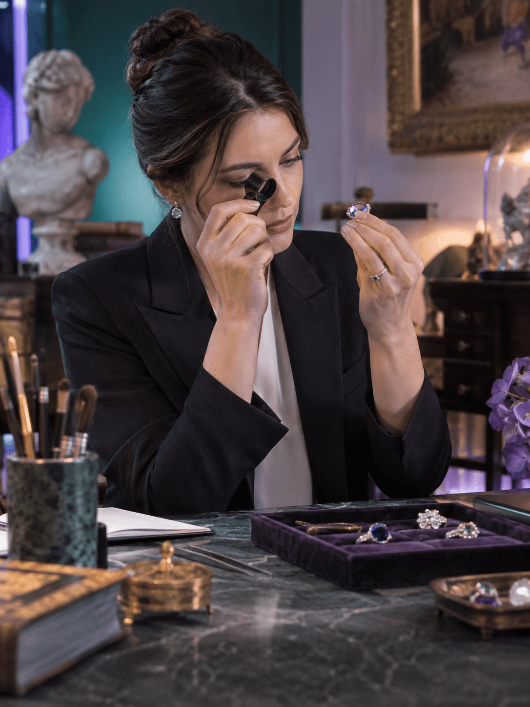 A jewelry catalog specialist using a loupe to inspect a gemstone at a softly lit expert workstation